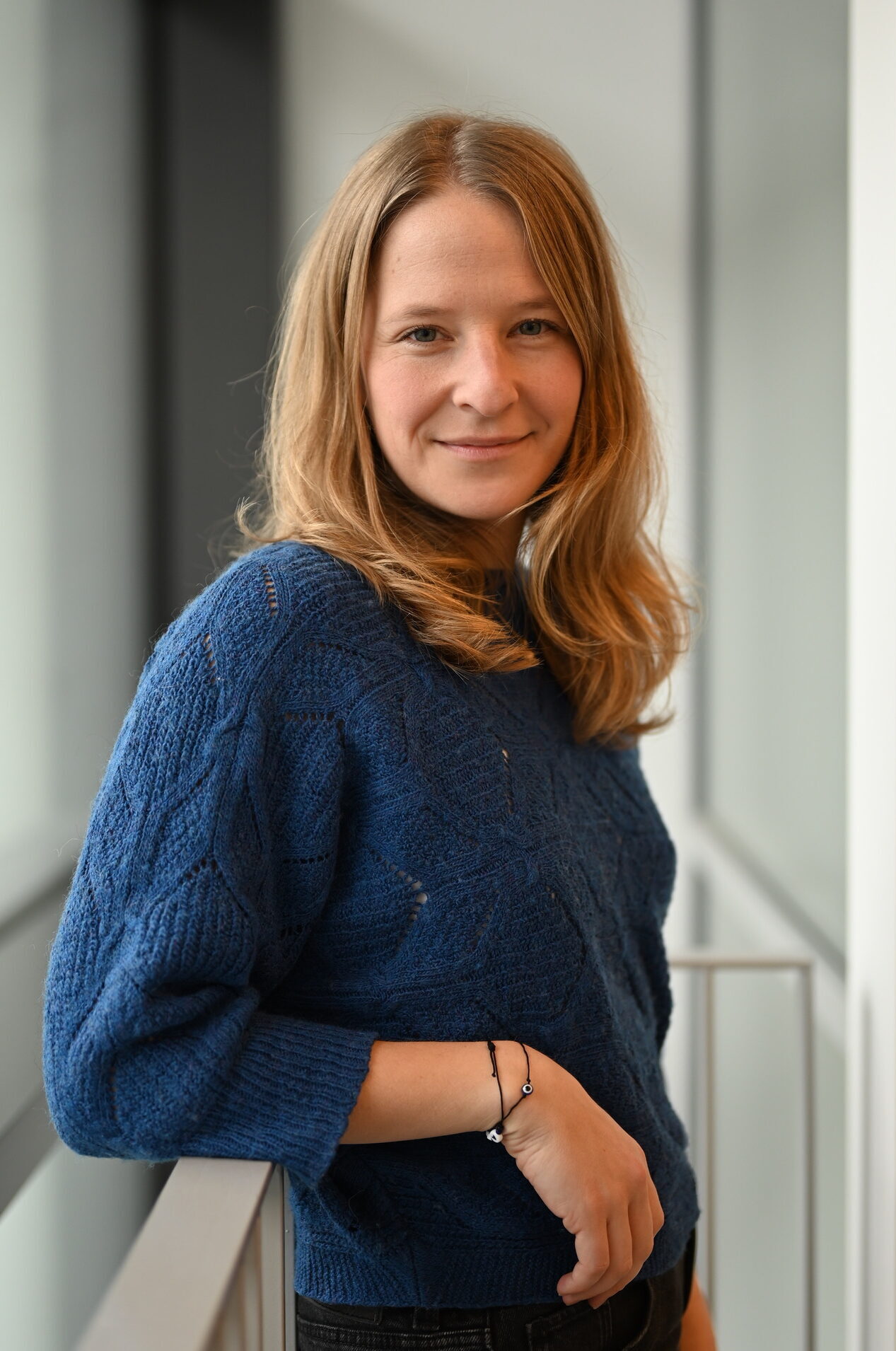 Das Bild zeigt mich, ich trage einen blauen Strickpulli und lehne mich an ein Geländer in einem Hausflur